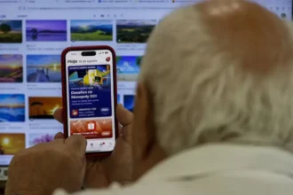 Imagem mostra idoso usando celular em frente a tela do computador