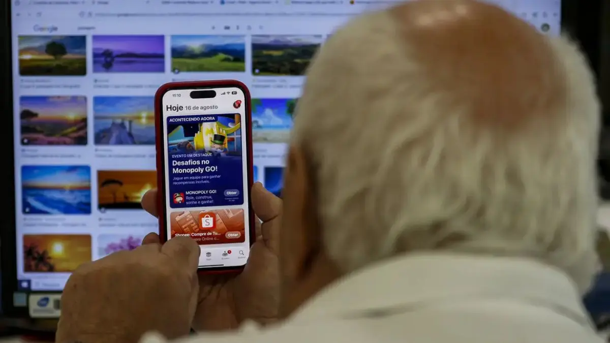 Imagem mostra idoso usando celular em frente a tela do computador